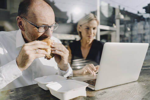 Businessman eating hamburger in coffee shop, while colleague is using laptop - KNSF06379