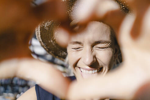 Woman with straw hat, looking into the sun, making heart shaped finger frame - KNSF06382