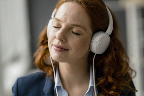 Portrait of redheaded businesswoman listening music with white headphones - KNSF06437