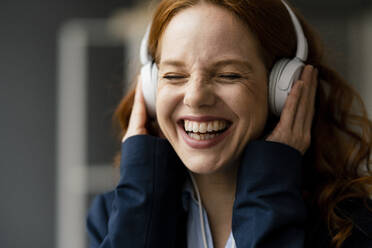 Portrait of laughing redheaded businesswoman listening music with white headphones - KNSF06439