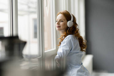 Portrait of redheaded businesswoman listening music with headphones while looking out of window - KNSF06440