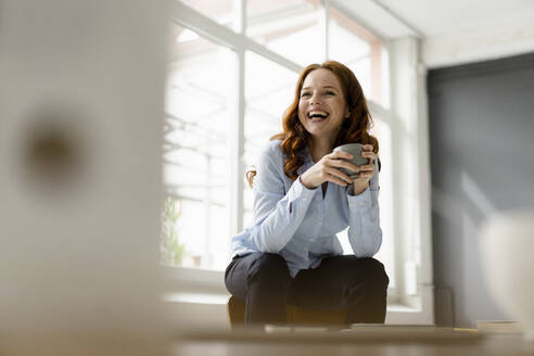 Portrait of laughing redheaded woman with tea bowl sitting on backrest in a loft - KNSF06445