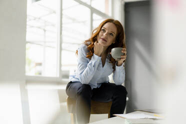 Portrait of pensive redheaded woman with tea bowl sitting on backrest in a loft - KNSF06446