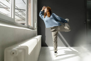 Redheaded woman standing on one leg in a loft looking out of window - KNSF06453