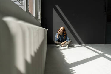 Rothaarige Frau sitzt auf dem Boden eines Dachbodens und genießt das Sonnenlicht - KNSF06454