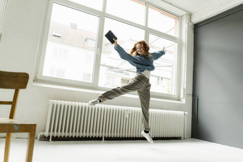 Porträt einer rothaarigen Frau mit digitalem Tablet, die auf einem Dachboden in die Luft springt, lizenzfreies Stockfoto