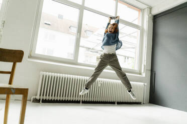 Portrait of screaming redheaded woman with digital tablet jumping in the air in a loft - KNSF06468