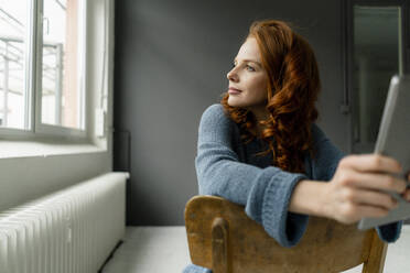 Portrait of redheaded woman with digital tablet sitting in a loft looking out of window - KNSF06469