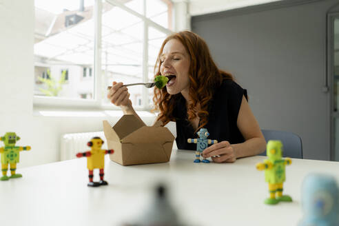 Portrait of redheaded businesswoman in a loft with miniature robots on desk eating healthy takeaway food - KNSF06485