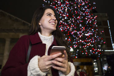 Portrait of happy young woman with smartphone in front of lighted Christmas tree outdoors - ABZF02573