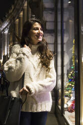 Porträt einer glücklichen jungen Frau mit Strickpullover, die in ein Schaufenster zur Weihnachtszeit schaut - ABZF02584