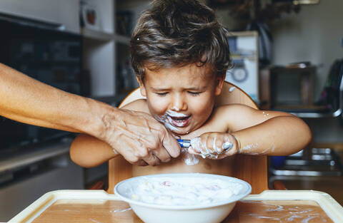Kleiner Junge isst Joghurt zu Hause, Hand der Frau auf dem Löffel, lizenzfreies Stockfoto