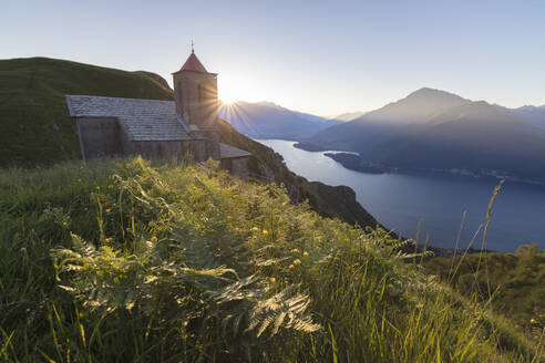 Sonnenstrahl auf der Kirche von San Bernardo beleuchtet die Landschaft um das blaue Wasser des Comer Sees in der Morgendämmerung, Musso, Lombardei, Italien, Europa - RHPLF08795