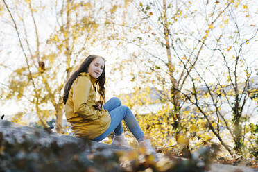 Mädchen in gelbem Regenmantel sitzt bei Bäumen - FOLF10949