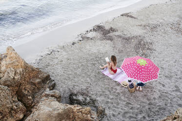 Junge Frau sitzt auf einem Handtuch am Strand und liest ein Buch - IGGF01305