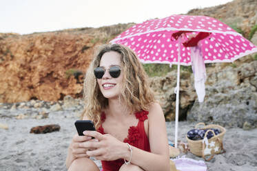 Lächelnde junge Frau am Strand sitzend mit Mobiltelefon - IGGF01309