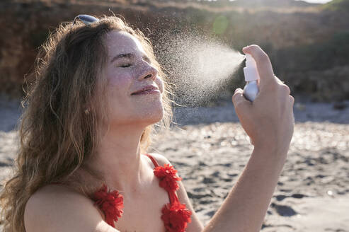 Young woman applying suncream spray on the beach - IGGF01320