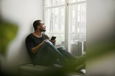 Mittelgroßer erwachsener Mann, der ein Smartphone auf dem Fensterbrett benutzt - FOLF11388