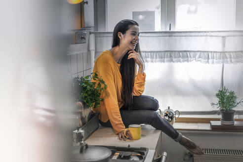 Happy young woman sitting on kitchen counter at home - RIBF00983