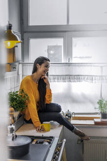 Happy young woman sitting on kitchen counter at home - RIBF00984