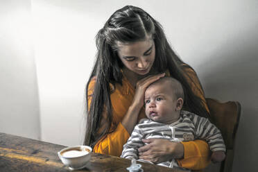 Mutter mit Baby sitzt am Holztisch zu Hause - RIBF00990