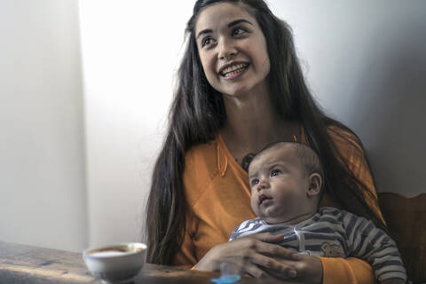 Glückliche Mutter mit Baby sitzt am Holztisch zu Hause, lizenzfreies Stockfoto