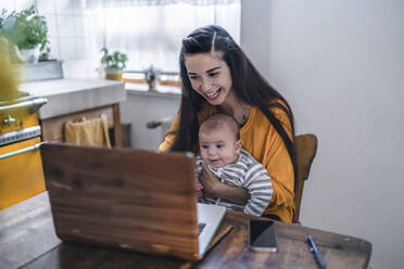 Laughing mother with baby using laptop on kitchen table - RIBF00994