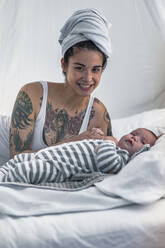 Porträt einer lächelnden, tätowierten jungen Frau mit ihrem Baby im Himmelbett - RIBF00997