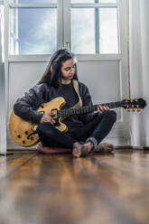 Young woman sitting on the floor at home playing guitar - RIBF01019