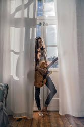 Serious young woman standing at the window at home with a guitar - RIBF01020