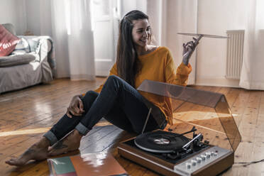 Smiling young woman sitting on the floor at home with record and record player - RIBF01024