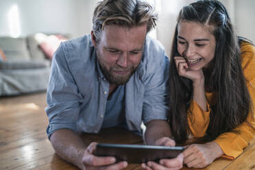 Happy couple lying on the floor at home looking at tablet - RIBF01037