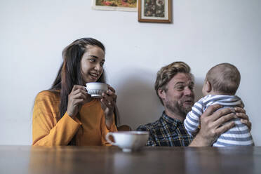 Glückliche Familie mit Baby sitzt am Holztisch zu Hause - RIBF01043
