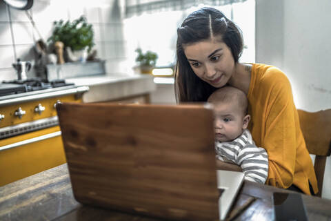 Mutter mit Baby am Laptop auf dem Küchentisch, lizenzfreies Stockfoto