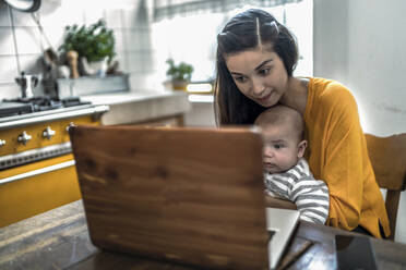 Mother with baby using laptop on kitchen table - RIBF01048