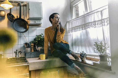 Young woman sitting on kitchen counter at home looking out of window stock photo