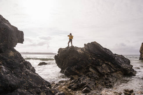 Woman wearing yellow rain jacket standing at rocky beach - UUF18965