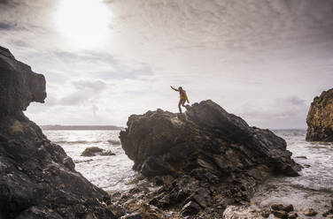 Woman wearing yellow rain jacket standing at rocky beach - UUF18967