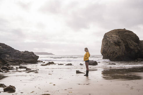 Woman wearing yellow rain jacket standing at rocky beach - UUF18974