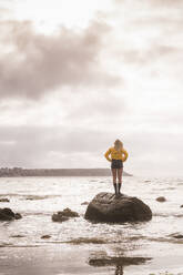 Woman wearing yellow rain jacket standing on rock - UUF18975