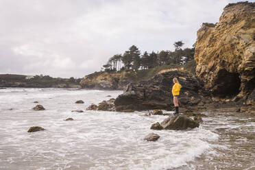 Woman wearing yellow rain jacket standing on rock at the beach - UUF18978