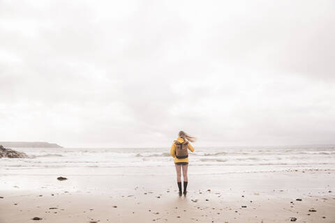 Rückansicht einer Frau mit gelber Regenjacke, die am Strand steht, lizenzfreies Stockfoto