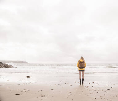 Rückansicht einer Frau mit gelber Regenjacke, die am Strand steht - UUF18982