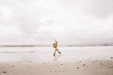 Woman wearing yellow rain jacket walking at the beach - UUF18983