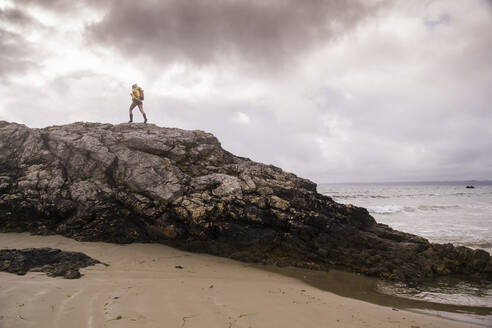 Woman wearing yellow rain jacket standing at rocky beach - UUF18987