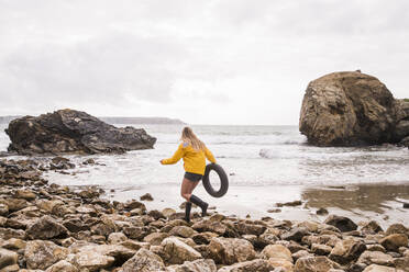 Frau in gelber Regenjacke mit Schwimmreifen, die an einem felsigen Strand spazieren geht - UUF18988