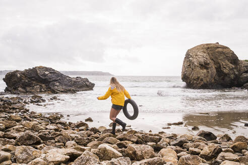 Woman wearing yellow rain jacket with floating tire, walking aon rocky beach - UUF18988