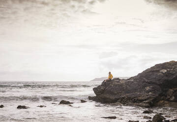 Woman wearing yellow rain jacket standing at rocky beach - UUF18993