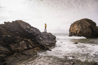 Woman wearing yellow rain jacket standing on rock at the beach - UUF18994