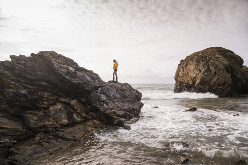 Woman wearing yellow rain jacket standing on rock at the beach - UUF18994